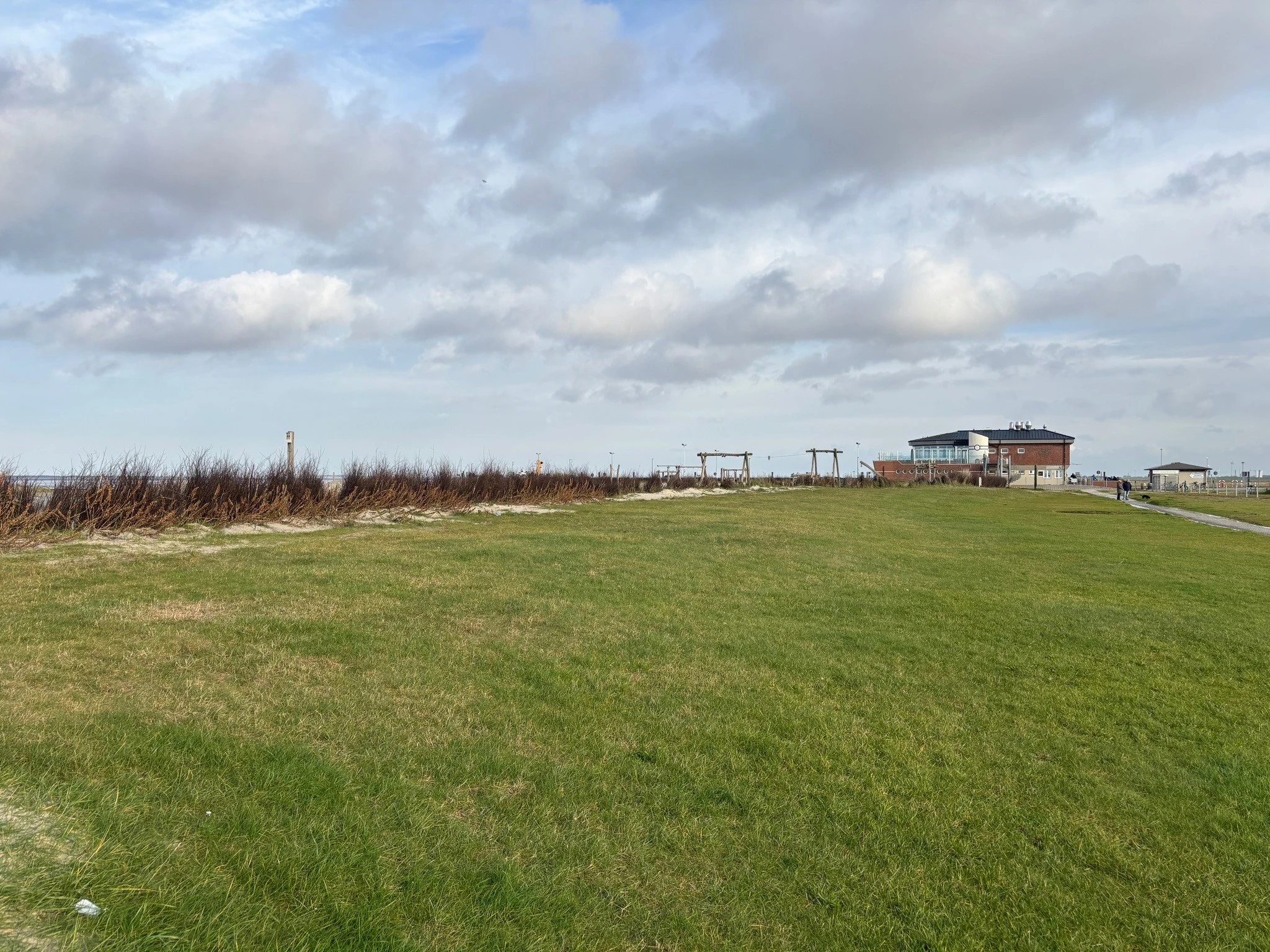 Großzügiger Grünstrand in Neßmersiel an der Nordsee mit Blick auf das Strandgebäude, den Kinderspielplatz und die Dünen unter bewölktem Himmel.