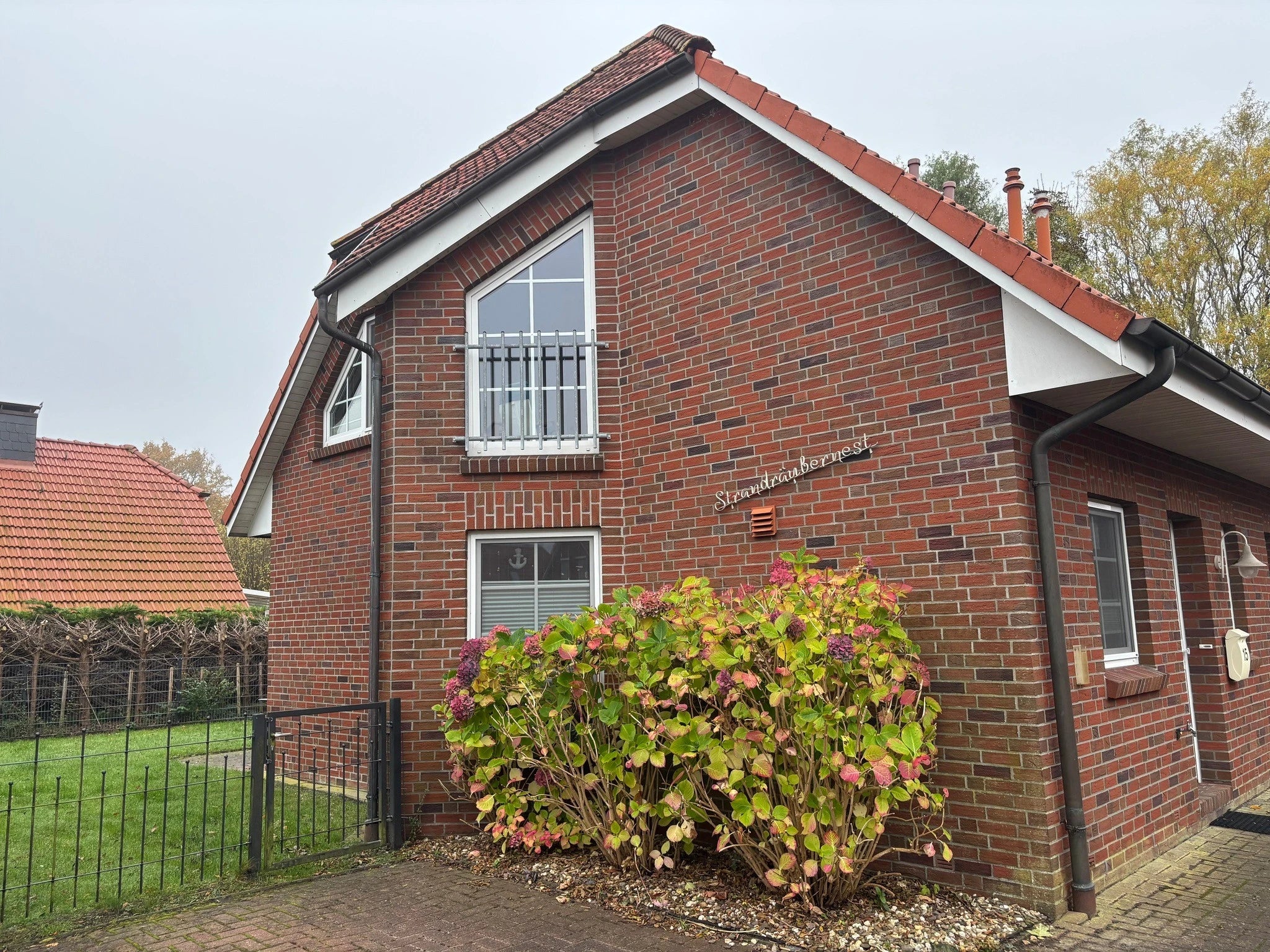 Rotes Klinker-Ferienhaus Strandräubernest in Neßmersiel an der Nordsee mit eingezäuntem Garten, Zaun und großem Hortensienbusch an der Hauswand.
