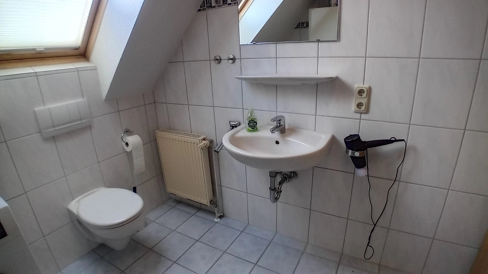 Helles Badezimmer im Obergeschoss des Ferienhauses Strandräubernest Neßmersiel mit weißem Waschbecken, Spiegel, Toilette und einem an der Wand hängenden Föhn unter der Dachschräge.