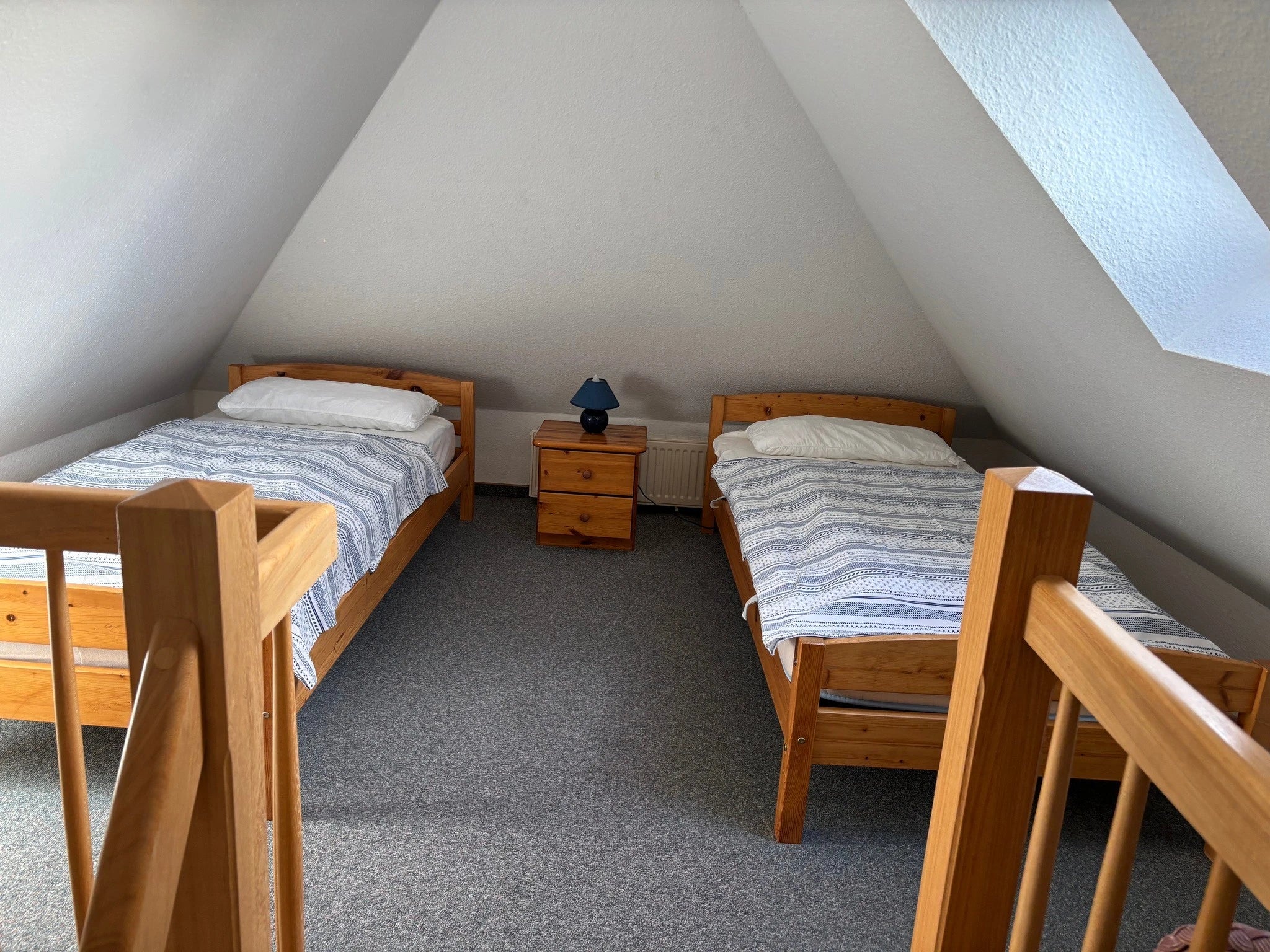Blick vom hölzernen Treppenaufgang in das zweite Schlafzimmer im Obergeschoss des Ferienhauses Strandräubernest Neßmersiel mit zwei Einzelbetten aus Holz und einem Nachttisch unter den Dachschrägen.