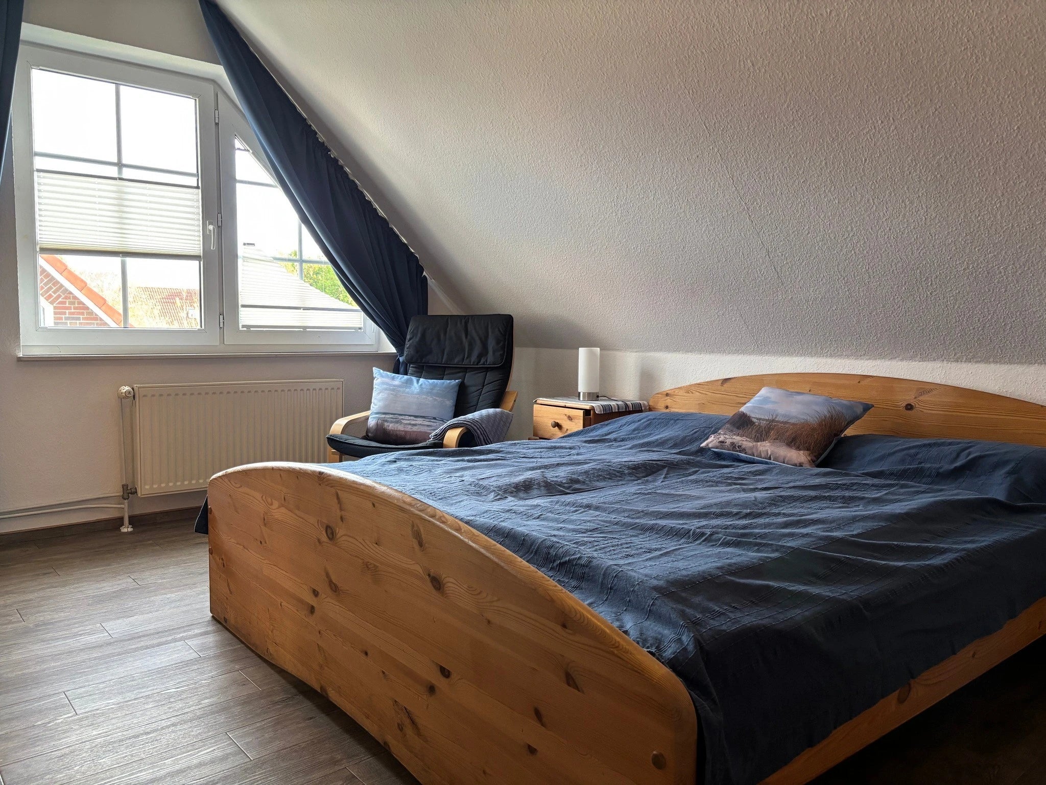 Gemütliches Elternschlafzimmer im Ferienhaus Strandräubernest Neßmersiel an der Nordsee mit großem Doppelbett aus Massivholz, Lesesessel und maritimen Kissen unter der Dachschräge.