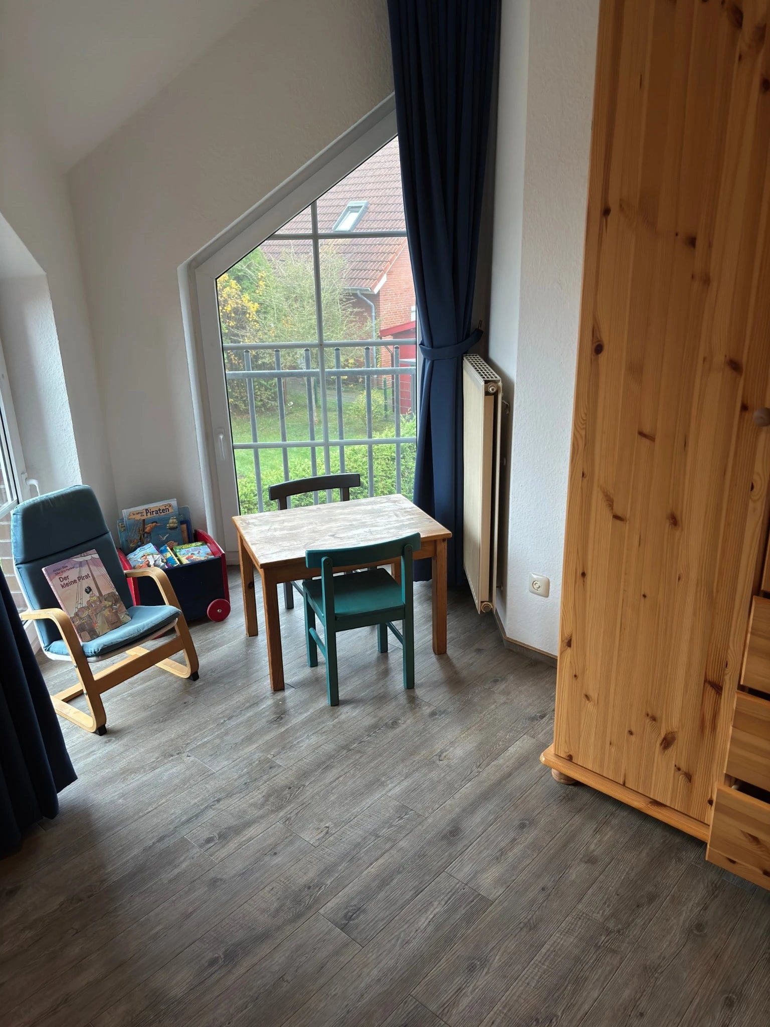 Familienfreundliche Kinderspielecke im Obergeschoss des Ferienhauses Strandräubernest Neßmersiel mit Kindertisch, Holzschrank, Sessel und einer Kiste voller Kinderbücher vor dem großen Giebelfenster.