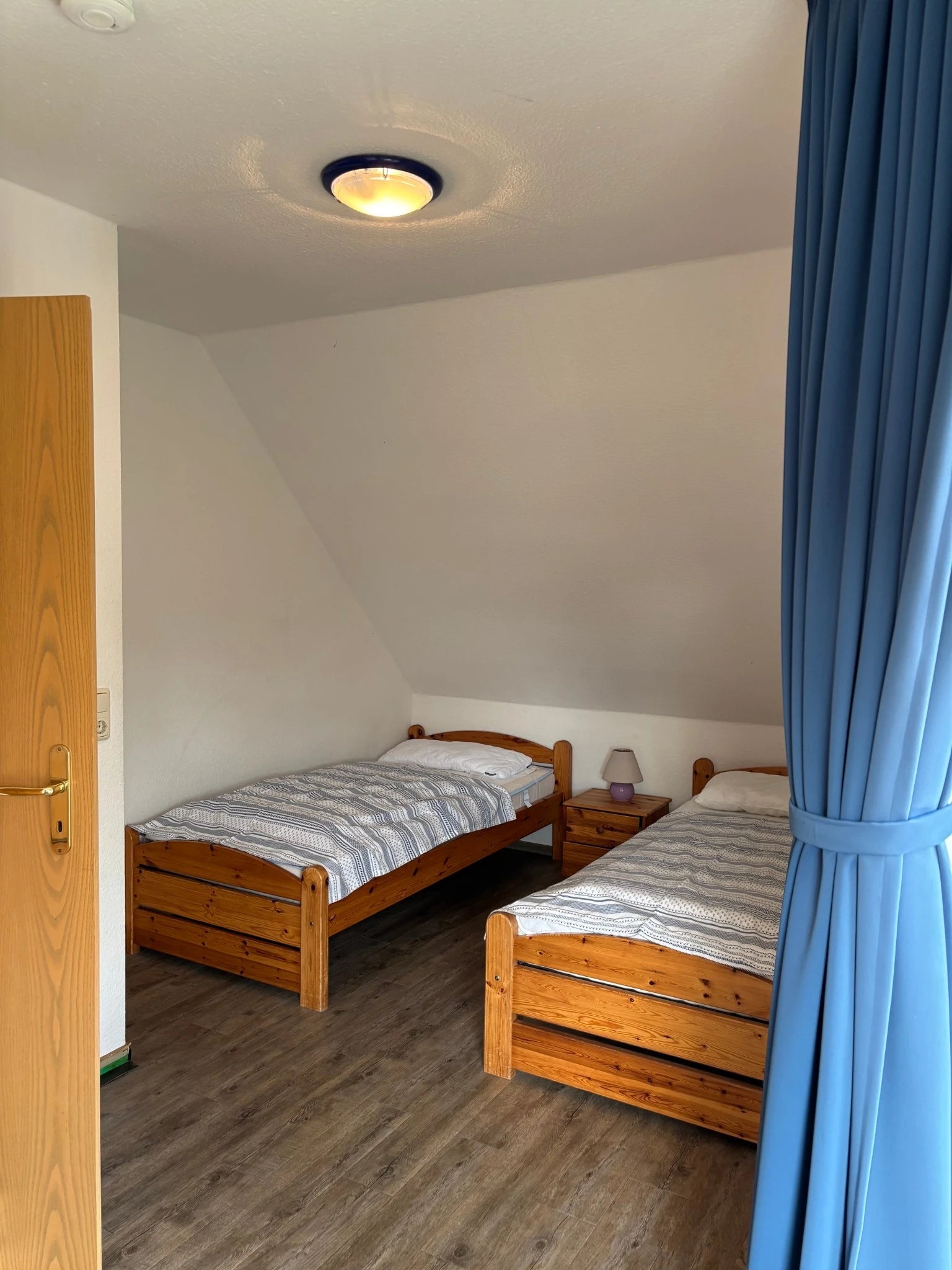 Gemütliches zweites Schlafzimmer im Obergeschoss des Ferienhauses Strandräubernest Neßmersiel mit zwei Einzelbetten aus Holz, Nachttisch und blauen Vorhängen unter der Dachschräge.