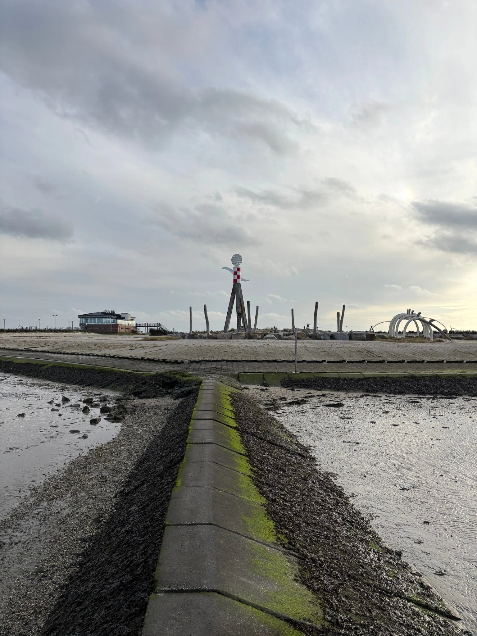 Blick über eine bemooste Steingalbuhne auf den Abenteuerspielplatz am Strand von Neßmersiel mit der rot-weißen Kletterbake und dem Strandgebäude im Hintergrund.